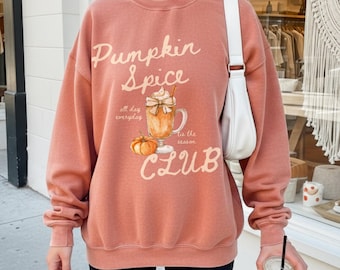 May include: Terracotta-colored sweatshirt with the text "Pumpkin Spice Club" and a graphic of a pumpkin spice latte and a pumpkin. The sweatshirt has long sleeves and a crew neck.