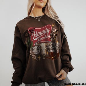May include: Dark chocolate brown sweatshirt with a graphic design. The design includes the words "Howdy Cowboy" in a red banner, a steer skull, a disco ball, a cactus, and a cowboy hat. The text "The Cowgirl Club" is also included.