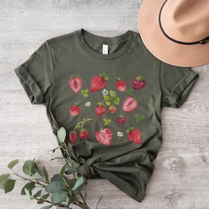 May include: A green t-shirt with a white crew neck and a red strawberry print. The shirt is laid flat on a wooden surface with a brown hat next to it.