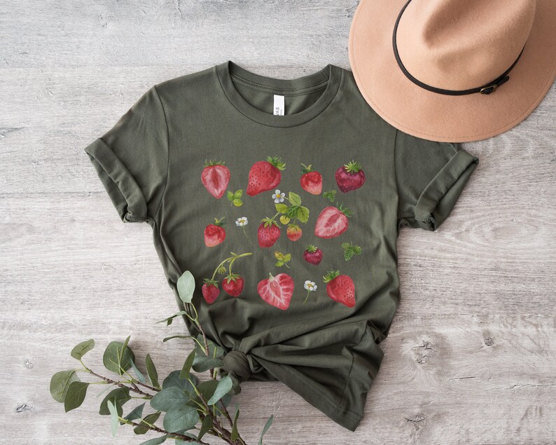 May include: A green t-shirt with a white crew neck and a red strawberry print. The shirt is laid flat on a wooden surface with a brown hat next to it.