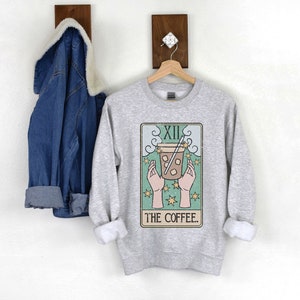 Puede incluir: Una sudadera gris claro con un dise&ntilde;o de carta del tarot que presenta una bebida de caf&eacute;. El dise&ntilde;o incluye el n&uacute;mero romano XII, manos sosteniendo una taza de caf&eacute; y las palabras "THE COFFEE". Una chaqueta vaquera cuelga de un gancho de pared de madera.