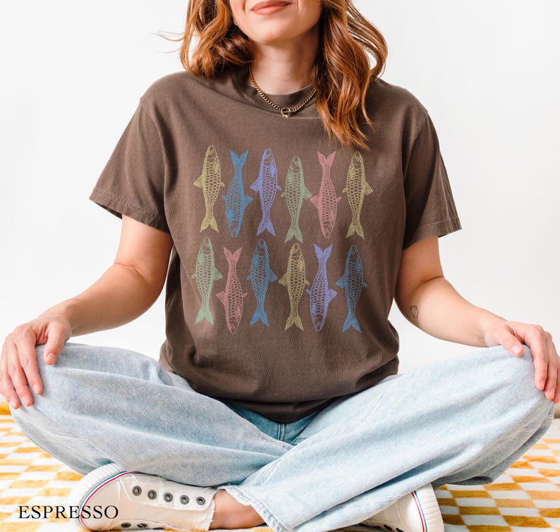 May include: A brown t-shirt with a colorful fish pattern. The shirt is being worn by a person with long brown hair. The person is sitting with their legs crossed and wearing light blue jeans.