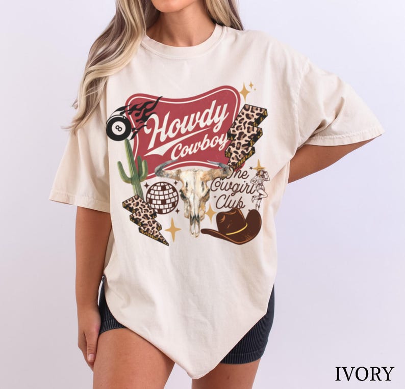 May include: Ivory-colored t-shirt with a graphic design featuring a steer skull, cowboy hat, and the words "Howdy Cowboy" and "The Cowgirl Club." The design includes a cactus, lightning bolt, and disco ball.