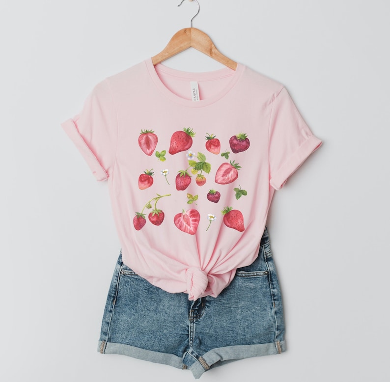 May include: A pink t-shirt with a graphic print of red strawberries and white flowers.