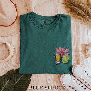 May include: A teal green t-shirt with a graphic of a shot glass of tequila, a lime wedge, and a pink flower.