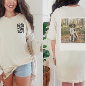 May include: A cream-coloured t-shirt with the text "Cool Dog Moms Club" printed on the front. The back of the shirt features a photo of a brown and white dog in a woodland setting.