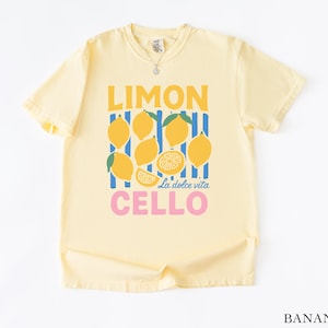 May include: A light yellow t-shirt with a graphic design featuring lemons and the words "LIMON CELLO" in yellow and pink. The design includes the phrase "La dolce vita". The shirt is made of a soft material.