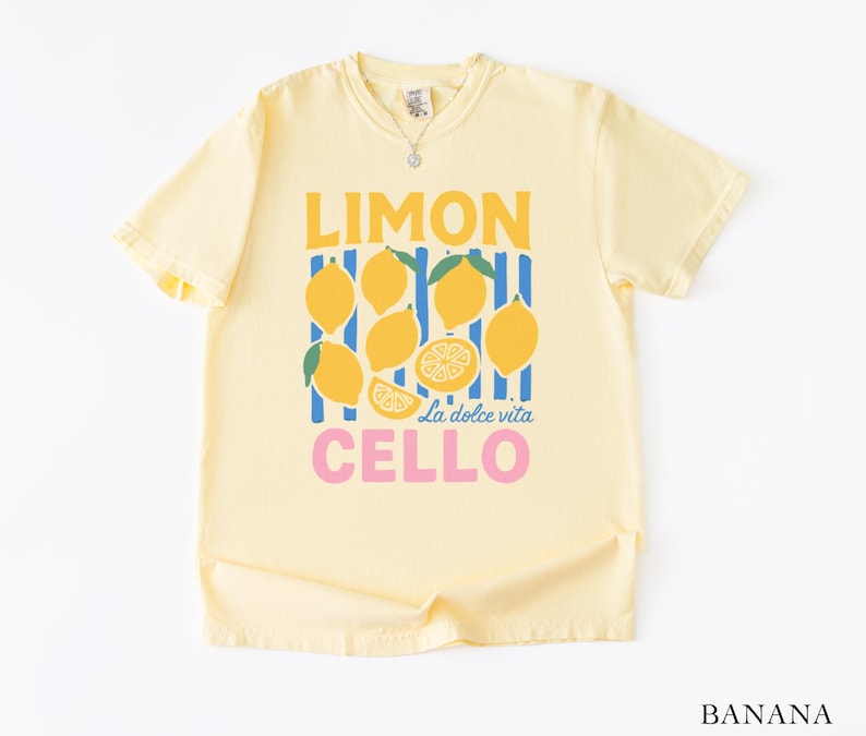 May include: A light yellow t-shirt with a graphic design featuring lemons and the words "LIMON CELLO" in yellow and pink. The design includes the phrase "La dolce vita". The shirt is made of a soft material.