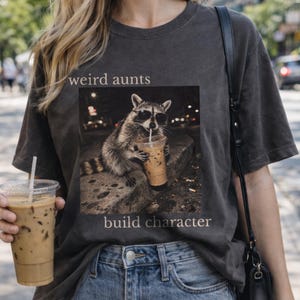 May include: Dark grey t-shirt with a raccoon in sunglasses drinking iced coffee, text reads "weird aunts build character." The raccoon graphic is in a square, with a blurred city background. The shirt is worn with blue jeans.