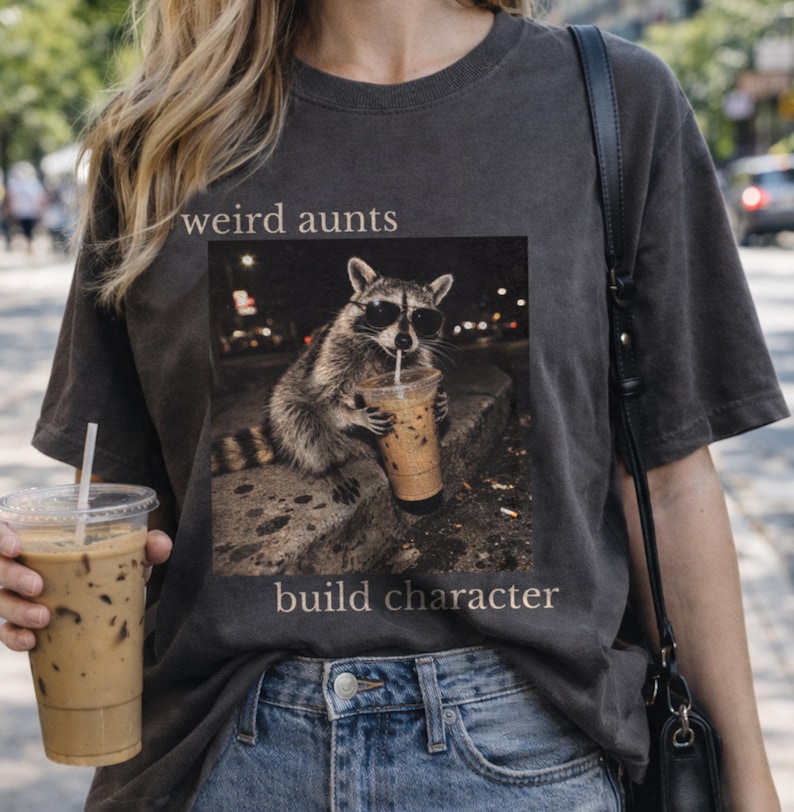 May include: Dark grey t-shirt with a raccoon in sunglasses drinking iced coffee, text reads "weird aunts build character." The raccoon graphic is in a square, with a blurred city background. The shirt is worn with blue jeans.