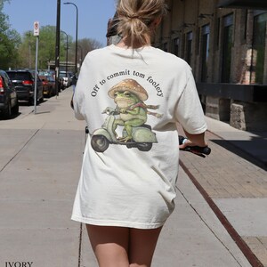 Peut inclure: Un t-shirt blanc avec un graphique d'une grenouille conduisant un scooter. Le texte "Off to commit tom foolery" est imprimé au dos du t-shirt.
