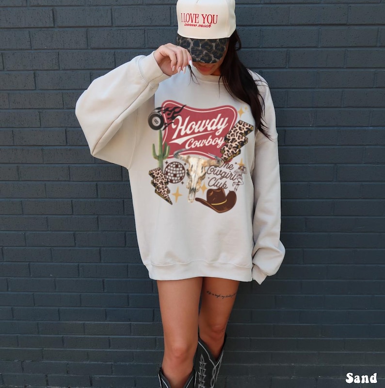 May include: Sand-colored sweatshirt with a western-themed graphic. The graphic includes the words "Howdy Cowboy" in a red banner, a steer skull, a cactus, and a cowboy hat. The sweatshirt is paired with a baseball cap that says "I love you" on the front.