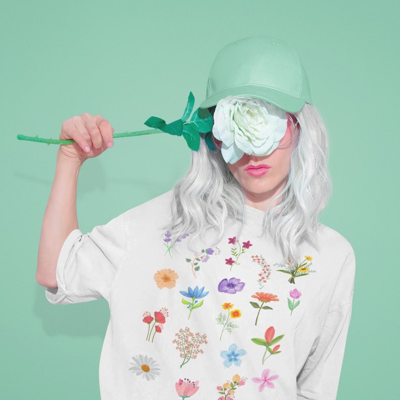 May include: A white t-shirt with a floral print featuring various flowers in different colors, including pink, blue, yellow, and red. The model is wearing a green baseball cap and holding a single white rose in front of their face.