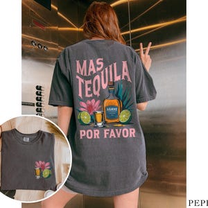 May include: A dark gray t-shirt with a graphic print of a tequila bottle, shot glass, and limes. The text on the shirt reads "Mas Tequila Por Favor".