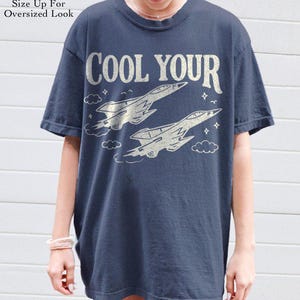 Puede incluir: Camiseta azul marino con el texto "COOL YOUR" sobre dos ilustraciones estilizadas de aviones de combate. La camiseta tiene cuello redondo y mangas cortas. El texto "Size Up For Oversized Look" est&aacute; impreso en la parte superior izquierda.