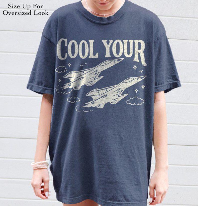 Puede incluir: Camiseta azul marino con el texto "COOL YOUR" sobre dos ilustraciones estilizadas de aviones de combate. La camiseta tiene cuello redondo y mangas cortas. El texto "Size Up For Oversized Look" est&aacute; impreso en la parte superior izquierda.