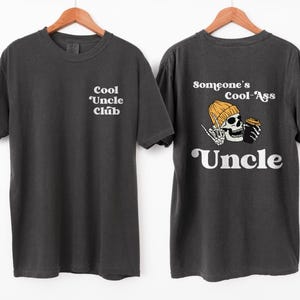 May include: Two black t-shirts with white text. The front shirt says "Cool Uncle Club". The back shirt says "Someone's Cool-Ass Uncle" with a cartoon skeleton wearing a yellow beanie and holding a cup of coffee.