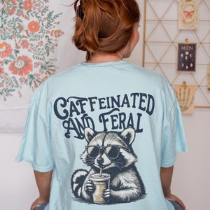 May include: A light blue t-shirt with a graphic of a raccoon wearing sunglasses and drinking a coffee. The text "caffeinated and feral" is printed on the back of the shirt.