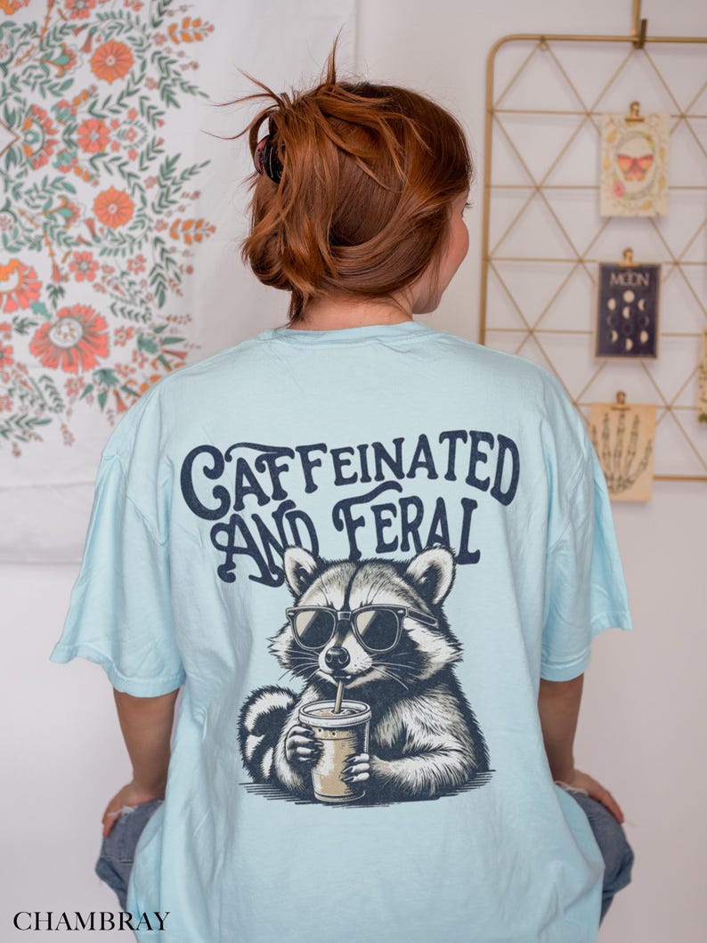 May include: A light blue t-shirt with a graphic of a raccoon wearing sunglasses and drinking a coffee. The text "caffeinated and feral" is printed on the back of the shirt.