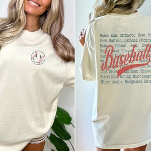 Könnte beinhalten: Ein cremefarbenes T-Shirt mit einem Baseball-Smiley auf der Vorderseite und einer Liste von Baseballbegriffen auf der Rückseite. Der Text auf der Rückseite lautet "Home Run. Strikeout. Base. In-field. Outfield. Diamond. Pitching. Batter. Fielder. Foul. Double. But Steal. Umpir. Curv ball. Baseball. Sacrifice. Line Drive and Slam. Shutout. Strike Zone. Inning. Major League. Minor League. Designated Hitter."