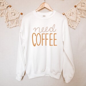 May include: White sweatshirt with the text "need coffee" in brown and pink lettering.