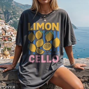 May include: Dark gray t-shirt with a lemon graphic. The design includes the word "LIMON" in yellow, lemons, blue stripes, and the word "CELLO" in pink. The phrase "la dolce vita" is also included.