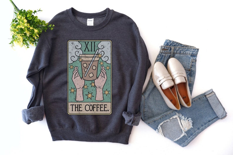 Puede incluir: Sudadera gris oscuro con un dise&ntilde;o de carta del tarot que representa una taza de caf&eacute;, el n&uacute;mero XII y las palabras "THE COFFEE". La sudadera se combina con vaqueros azules y mocasines blancos.