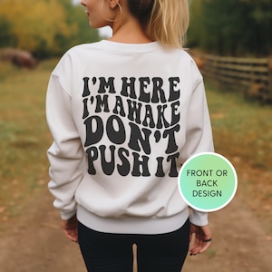 May include: White sweatshirt with black text that reads "I'm here I'm awake don't push it" in a retro font. The text is on the back of the sweatshirt. The sweatshirt is being worn by a person.