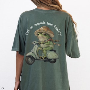 Puede incluir: Camiseta verde musgo con una ilustración caprichosa de una rana montando una scooter, con un sombrero de seta y una bufanda. El texto "Off to commit tom foolery" está arqueado sobre la rana. La camiseta es de corte holgado.