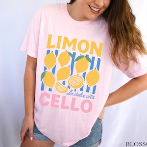 May include: A light pink t-shirt with a lemon-themed graphic. The design features the word "LIMON" in yellow, lemons, blue stripes, and the words "CELLO La dolce vita" in pink. The shirt is a casual style.