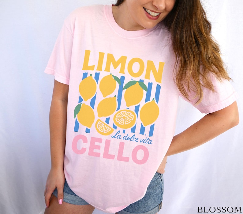 May include: A light pink t-shirt with a lemon-themed graphic. The design features the word "LIMON" in yellow, lemons, blue stripes, and the words "CELLO La dolce vita" in pink. The shirt is a casual style.