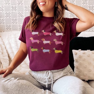 May include: A maroon t-shirt with a graphic print of nine dachshund dogs wearing pajamas. The dogs are in various colors and patterns.