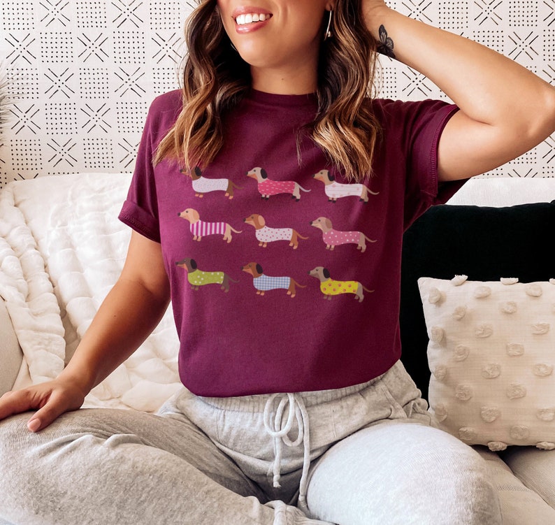 May include: A maroon t-shirt with a graphic print of nine dachshund dogs wearing pajamas. The dogs are in various colors and patterns.
