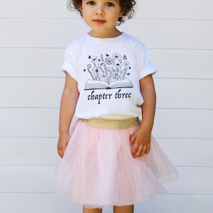 May include: A child's outfit featuring a white t-shirt with a black floral design and the text "chapter three." The outfit includes a pink tulle skirt with a gold waistband.