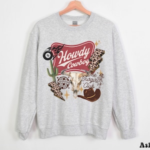 May include: A light grey sweatshirt with a graphic design featuring a red banner that reads "Howdy Cowboy". The design includes a bull skull, cactus, lightning bolts, and a cowboy hat. The text "The Cowgirl Club" is also present.