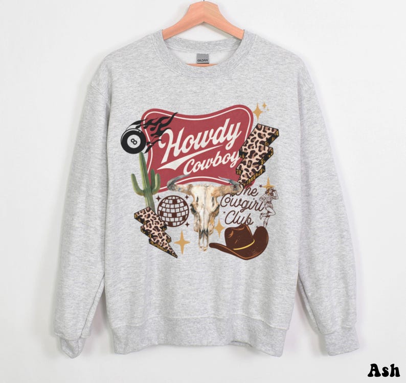 May include: A light grey sweatshirt with a graphic design featuring a red banner that reads "Howdy Cowboy". The design includes a bull skull, cactus, lightning bolts, and a cowboy hat. The text "The Cowgirl Club" is also present.