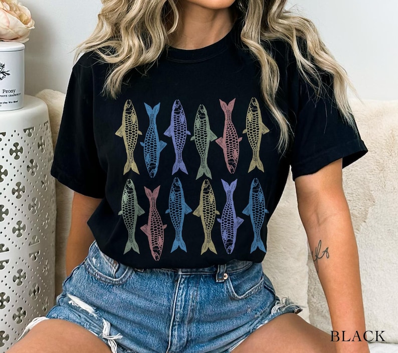 May include: A black t-shirt with a colorful fish pattern. The fish are arranged in rows and are outlined in black. The shirt is being worn by a person with long blonde hair.
