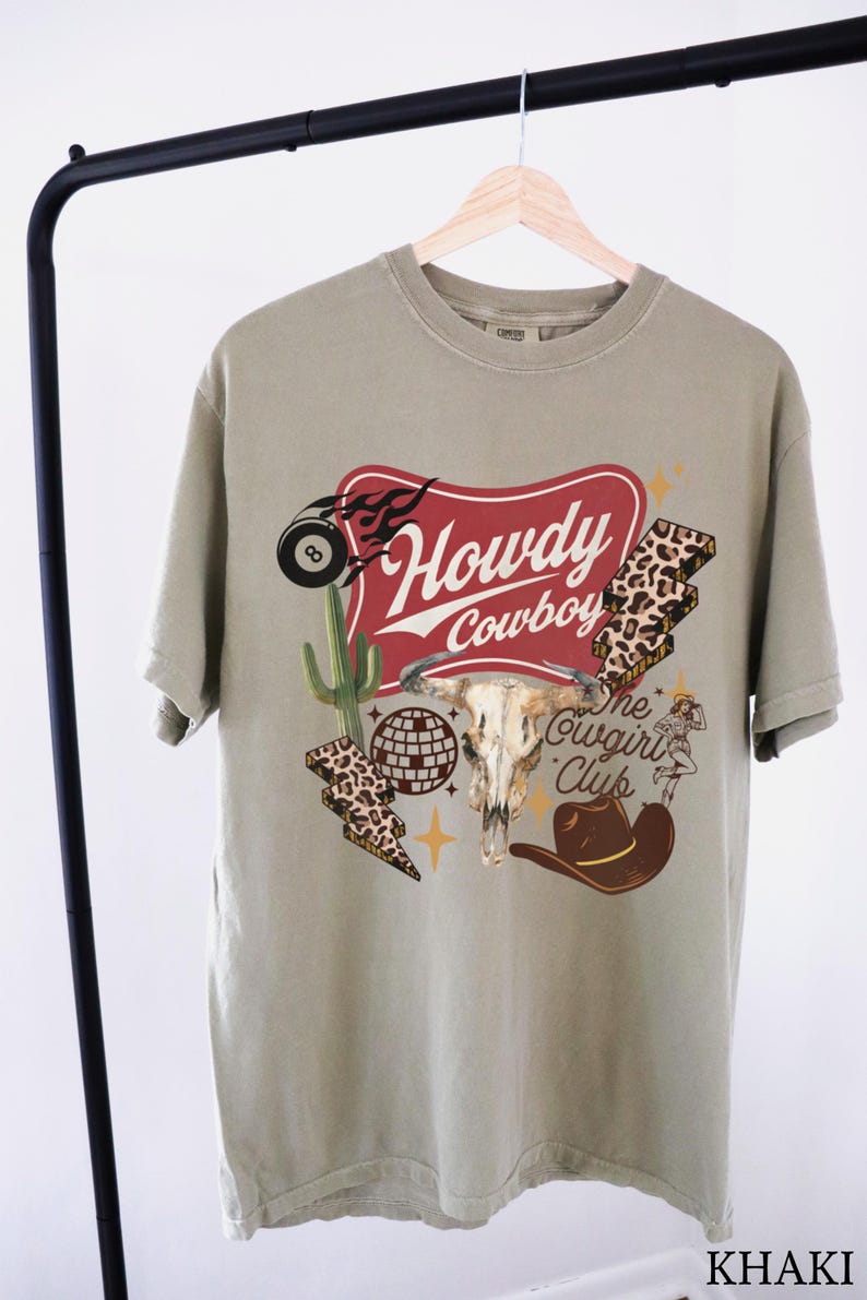 May include: Khaki-colored t-shirt with a graphic design featuring a steer skull, cowboy hat, and the words "Howdy Cowboy" in a red banner. Additional graphics include a cactus, lightning bolts, and an 8-ball.