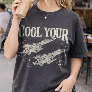 Puede incluir: Camiseta gris oscuro con el texto "COOL YOUR" sobre dos ilustraciones estilizadas de aviones de combate. El dise&ntilde;o incluye nubes y estrellas. La camiseta la lleva una persona que sostiene una bebida.