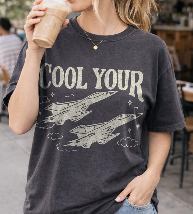 Puede incluir: Camiseta gris oscuro con el texto "COOL YOUR" sobre dos ilustraciones estilizadas de aviones de combate. El dise&ntilde;o incluye nubes y estrellas. La camiseta la lleva una persona que sostiene una bebida.