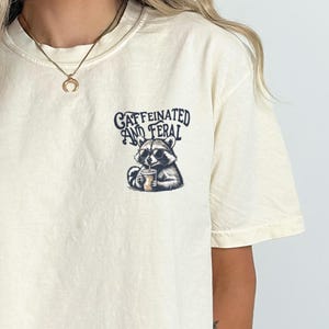 May include: A cream colored t-shirt with a graphic of a raccoon drinking from a cup. The text "caffeinated and feral" is printed above the raccoon.