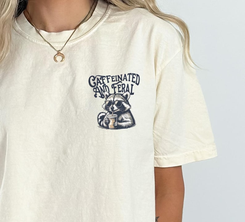 May include: A cream colored t-shirt with a graphic of a raccoon drinking from a cup. The text "caffeinated and feral" is printed above the raccoon.