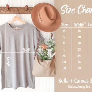 May include: Size chart for a unisex jersey tee, showing measurements in inches for width and length. The chart includes sizes XS through 3XL. The image also shows a gray t-shirt hanging on a wooden hanger, a brown hat, and a brown bag.