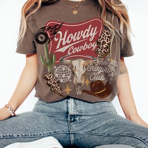 May include: Espresso-colored t-shirt with a western-themed graphic. The design includes a bull skull, cowboy hat, cactus, and the words "Howdy Cowboy" and "The Cowgirl Club". The shirt has a vintage, distressed look.
