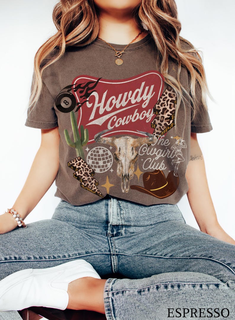 May include: Espresso-colored t-shirt with a western-themed graphic. The design includes a bull skull, cowboy hat, cactus, and the words "Howdy Cowboy" and "The Cowgirl Club". The shirt has a vintage, distressed look.