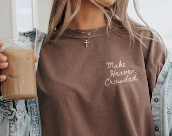 Op de afbeelding: Een bruin t-shirt met de geborduurde tekst "Make Heaven Crowded" in witte letters. De persoon draagt een zilveren kruis ketting en een spijkerjasje. Een helder glas met een bruine drank wordt in de hand gehouden.