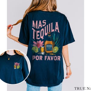 May include: A navy blue t-shirt with a graphic of a tequila bottle, shot glass, and flowers. The text "Mas Tequila Por Favor" is printed on the shirt.