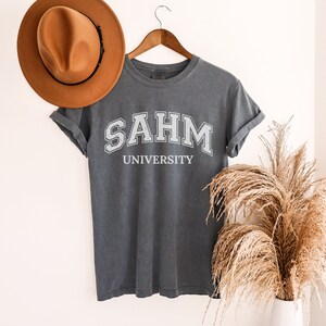 Puede incluir: Una camiseta gris oscura con el texto "SAHM UNIVERSITY" impreso en blanco en la parte delantera.