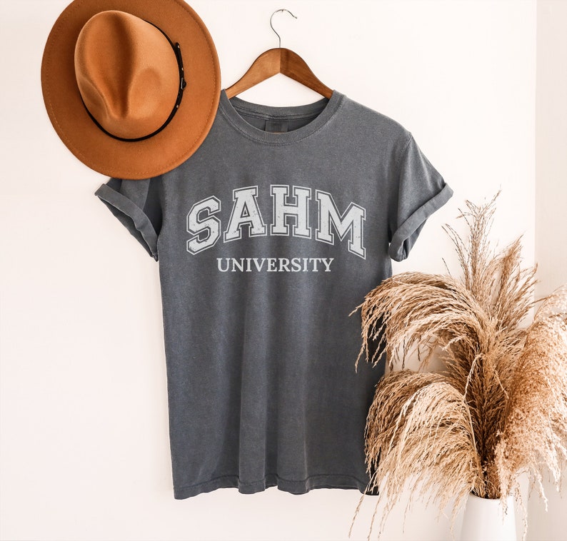 Puede incluir: Una camiseta gris oscura con el texto "SAHM UNIVERSITY" impreso en blanco en la parte delantera.