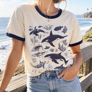 May include: A cream-colored ringer t-shirt with dark blue trim on the collar and sleeves. The front features a detailed dark blue graphic print of multiple orca whales, various smaller fish, and coral reef elements scattered across the fabric.